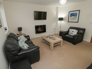 A living room with black leather sofas a wooden coffee table with cups and plates a wall-mounted tv and electric fireplace at Beach House in Beadnell