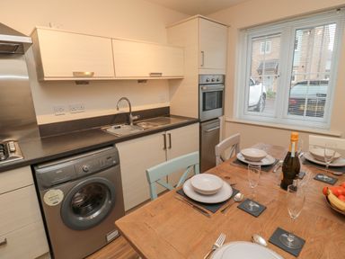 A kitchen with a washing machine a sink an oven a dining table set for four with plates glasses and a bottle at Beach House in Beadnell