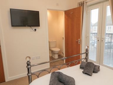 A bedroom with a metal bed frame folded towels a wall mounted television an open door leading to a bathroom and glass doors with blinds at Beach House in Beadnell
