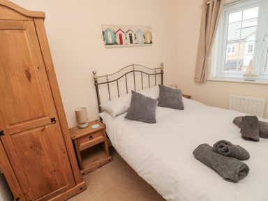 A bedroom with a double bed, two grey pillows, rolled towels, wooden wardrobe, nightstand with lamp, window with blinds, and wall art of beach huts at Beach House in Beadnell