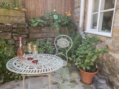 A small outdoor patio with a metal table and chair a bottle and two glasses on the table surrounded by plants at Crooked Cottage in Kirkbymoorside