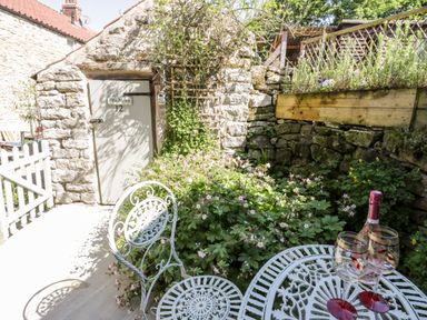 A stone courtyard with a white metal table and chairs wine bottle and glasses stone walls and plants at Crooked Cottage in Kirkbymoorside