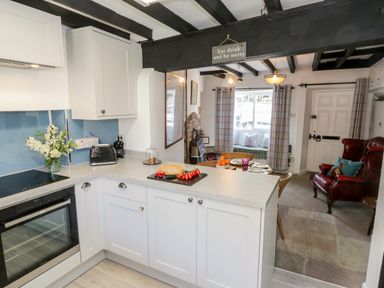 A kitchen with white cabinets and countertop adjacent to a dining area with a red armchair and a window seat at Crooked Cottage in Kirkbymoorside