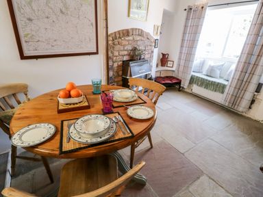 A dining area with a round wooden table set with plates glasses and cutlery a bowl of oranges a brick fireplace and a window seat with cushions at Crooked Cottage in Kirkbymoorside