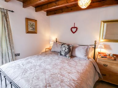 A bedroom with a bed a bedside table lamps a mirror a floral painting and a heart decoration at Crooked Cottage in Kirkbymoorside