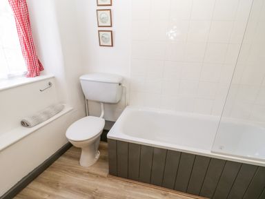 A bathroom with a toilet next to a bathtub and a window with red checkered curtains at Crooked Cottage in Kirkbymoorside