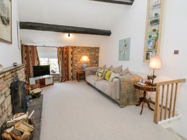 A living room with a beige sofa and cushions a small round table and lamp a TV near curtains and a fireplace at Crooked Cottage in Kirkbymoorside