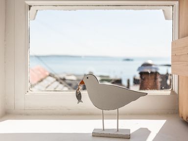 A seagull ornament holding a fish on a windowsill with a view of rooftops and the sea at Mole Cottage in Mousehole