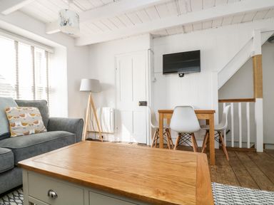 A living room with a wooden coffee table gray sofa with a bird pillow floor lamp dining table with white chairs and a wall-mounted TV at Mole Cottage in Mousehole