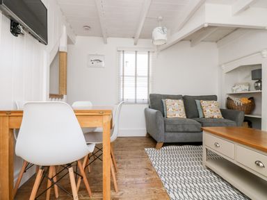 A living room with a gray sofa and pillows a wooden coffee table a dining table with white chairs and a wall mounted television at Mole Cottage in Mousehole