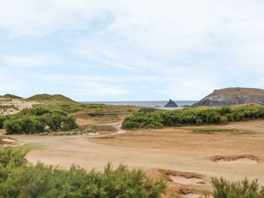 A coastal landscape with grassy dunes, sand patches, shrubs, sea in the background and rocky hills at Seaspell Beach House in Constantine Bay