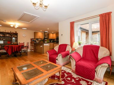 A living room with two armchairs covered with red throws a wooden coffee table and a dining table with chairs next to a kitchen area at Vine Lodge in Kingsbury Episcopi near Martock