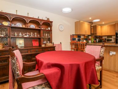A dining area with wooden chairs and a round table covered in a red tablecloth next to a wooden cabinet and kitchen at Vine Lodge in Kingsbury Episcopi near Martock