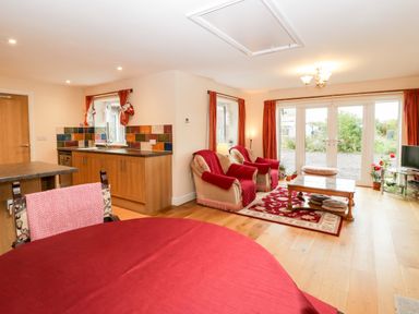 An open plan living room and kitchen with red tablecloth on dining table beige armchairs with red covers and a coffee table at Vine Lodge Kingsbury Episcopi near Martock