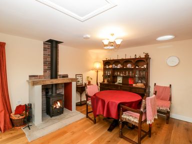 A dining room with a round table covered with a red tablecloth four chairs a wood burning stove and a wooden cabinet at Vine Lodge Kingsbury Episcopi near Martock