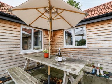 A wooden picnic table with an umbrella bottle of wine and two glasses outside a wooden building with tiled roof at Vine Lodge in Kingsbury Episcopi near Martock