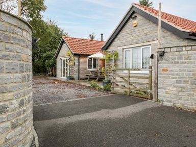 A stone and wood exterior of two single story buildings with tiled roofs and a wooden gate at Vine Lodge in Kingsbury Episcopi near Martock