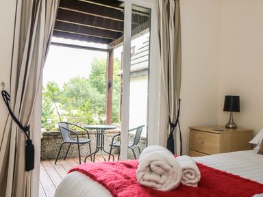 A bedroom with a bed and rolled towels next to a bedside table with a lamp and an open door leading to a balcony with a table and two chairs at Valley Lodge 48 in Callington