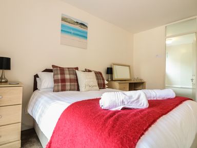 A bedroom with a double bed with pillows and towels a red blanket a wooden bedside table with a lamp a dressing table with a mirror and lamp a painting on the wall and a bathroom door at Valley Lodge 48 Callington