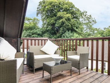 An outdoor wooden deck with wicker chairs and a glass top table surrounded by trees at Valley Lodge 48 in Callington