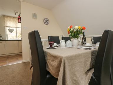 A dining area with a table covered by a beige tablecloth and set with dishes flowers and a jar of jam in a room with leather chairs and a clock on the wall at Valley Lodge 48 in Callington