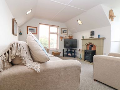 A living room with beige sofas a television on a stand a fireplace and framed pictures on the wall at The Watch in Porthcurno
