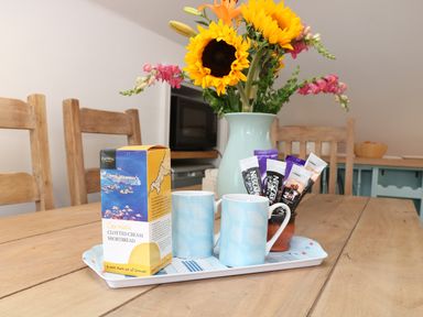 A wooden dining table with a tray holding two mugs, Nescafe sachets, and a box of Cornish clotted cream shortbread at The Watch in Porthcurno