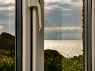 A window with a handle overlooking greenery and a body of water under a cloudy sky at The Watch in Porthcurno