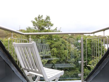 A white chair on a balcony with metal railing and glass panels overlooking trees at The Watch in Porthcurno