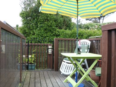 An outdoor patio with a green folding table under a yellow and green striped umbrella two white chairs and a bottle with two glasses at The Watch in Porthcurno