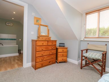 A bedroom corner with wooden dresser and chair next to a window and an open door showing a bathroom at The Watch in Porthcurno