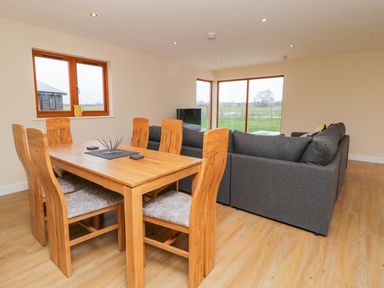 A dining area with a wooden table and six chairs next to a living room with a gray sofa and television at Oak Lodge in Sheriff Hutton