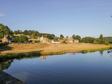 A river in front of dry grass fields with old stone ruins and houses in the background at Oak Lodge in Sheriff Hutton