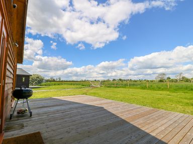 A wooden deck with a charcoal grill next to a grassy field with a wooden gate and fence at Oak Lodge in Sheriff Hutton