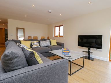 A living room with grey sofas and yellow patterned pillows a coffee table with board games a wall-mounted TV and wooden flooring at Rose Lodge in Sheriff Hutton