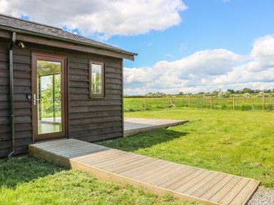 A wooden house with a glass door and window next to a wooden pathway and deck in a grassy field at Rose Lodge in Sheriff Hutton