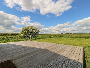 A wooden deck with grass and a large bush beyond a wire fence under a partly cloudy sky at Rose Lodge in Sheriff Hutton