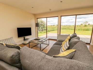 A living room with a grey sofa and patterned cushions a coffee table a television and large windows overlooking a grassy outdoor area at Rose Lodge in Sheriff Hutton