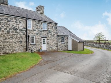 Exterior view of a stone-built house with white doors and windows near a paved driveway and green grass at Gwrtheyrn in Dyffryn Ardudwy