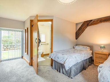 A bedroom with two single beds a wooden bedside table with a lamp an open door to a bathroom and a sliding glass door with vertical blinds at Gwrtheyrn in Dyffryn Ardudwy