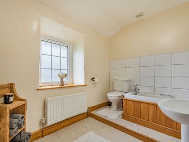 A bathroom with a bathtub, toilet, sink, wooden shelves with towels, and a window with a flower vase at Gwrtheyrn in Dyffryn Ardudwy