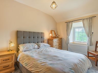 A bedroom with a double bed and patterned bedding a wooden bedside table with a lamp a wooden chest of drawers with a lamp and mirror wooden chair and window with light curtains at Gwrtheyrn in Dyffryn Ardudwy