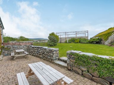 An outdoor patio with wooden picnic tables and stone walls overlooking a lawn with bushes and a metal frame structure at Gwrtheyrn in Dyffryn Ardudwy