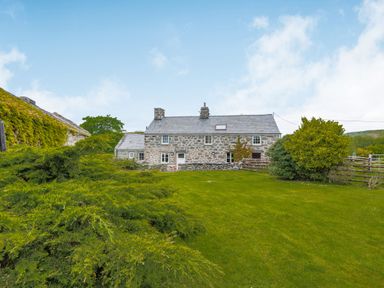 Stone house with slate roof surrounded by green lawn and bushes under blue sky at Gwrtheyrn in Dyffryn Ardudwy