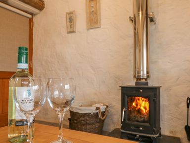 A wood burning stove with fire inside next to a basket and a table with a bottle of wine and two wine glasses at Edwin's View in Nancegollan nr Porthleven