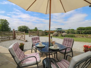 An outdoor patio with a table, four cushioned chairs, an umbrella, and a tractor in the background at Edwin's View in Nancegollan, nr Porthleven