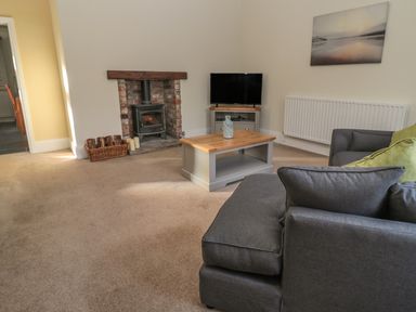 A living room with a gray sofa a wooden coffee table a TV on a stand a wood burning stove and a basket of firewood at Hare & Hounds House in Allendale