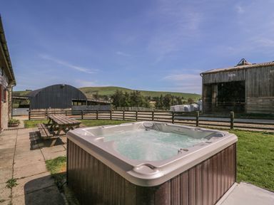 An outdoor hot tub with bubbling water beside a wooden picnic bench near farm buildings and green hills at Glanyrafon in St Harmon