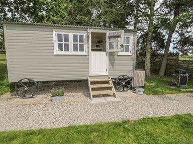 A small gray shepherd hut with wheels stairs and open door outside on a gravel path with a barbecue grill and trees at Bellwether in Helmsley