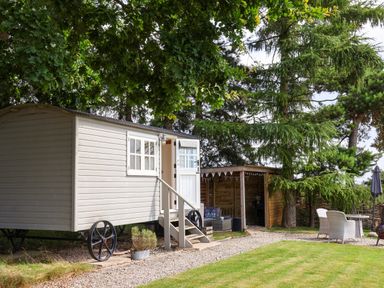 A small wagon-style cabin with stairs leading to an open door beside a wooden shelter with seating and a table with chairs in a garden at Bellwether in Helmsley